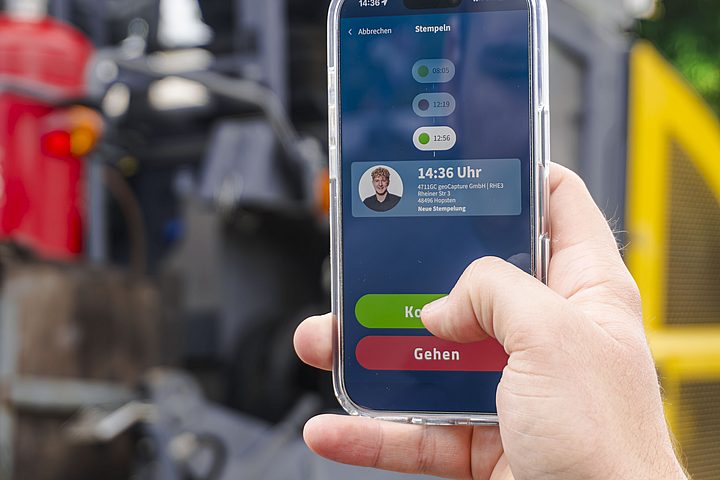 Eine Hand hält ein Smartphone mit geöffneter Zeiterfassungs-App. Auf dem Bildschirm sind ein Mitarbeiterprofil sowie grüne und rote Schaltflächen zum Starten und Beenden der Arbeitszeit zu sehen.