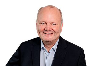 Porträt eines lächelnden Mannes mit kurzem grauem Haar, hellblauem Hemd und dunklem Sakko vor weißem Hintergrund.