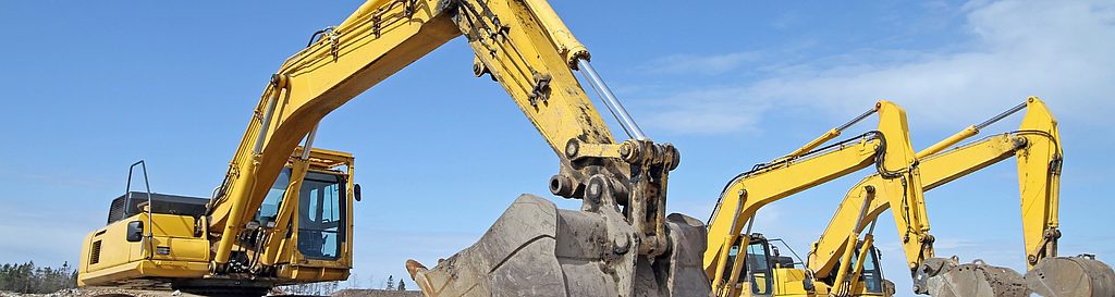 Mehrere gelbe Kettenbagger stehen auf einer Schotterfläche unter blauem Himmel; im Vordergrund ist die große Baggerschaufel einer Maschine zu sehen.