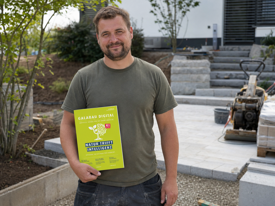 Ein Mann steht auf einer Baustelle im Garten- und Landschaftsbau und hält eine grüne Broschüre mit der Aufschrift „GaLaBau Digital – Natur trifft Intelligenz“. Im Hintergrund sind Pflasterarbeiten, Treppenstufen und Baumaschinen zu sehen.