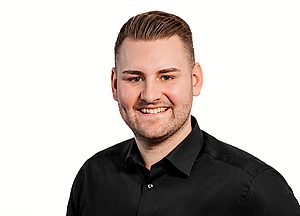 Porträt eines lächelnden jungen Mannes mit kurzen braunen Haaren, gepflegtem Bart und schwarzem Hemd vor weißem Hintergrund.