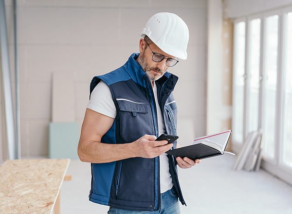 Mann mit Helm auf dem Kopf hält Handy und Notizbuch in den Händen