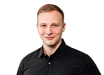 Porträtfoto eines lächelnden Mannes mit kurzem Haar, schwarzem Hemd und hellem Hintergrund