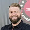 Porträt eines lächelnden Mannes mit Bart und tätowierten Unterarmen vor dem Logo „AGK Kampfmittelbergung“.
