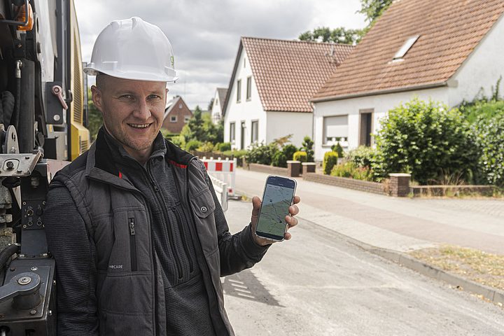 Mitarbeiter mit Schutzhelm zeigt die Fahrzeugortung auf einem Smartphone neben einem Fahrzeug.