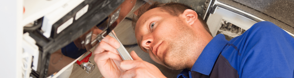 Ein Handwerker in blauer Arbeitskleidung arbeitet liegend unter einem technischen Gerät und zieht mit einem Schraubendreher eine Schraube fest