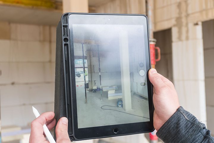 Eine Person hält auf einer Baustelle ein Tablet und dokumentiert damit den Innenraum eines Rohbaus. Auf dem Bildschirm ist die Kameraansicht des Raumes zu sehen.