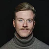 Porträt eines Mannes mit kurzem blondem Haar, Schnurrbart und grauem Rollkragenpullover vor dunklem Hintergrund.