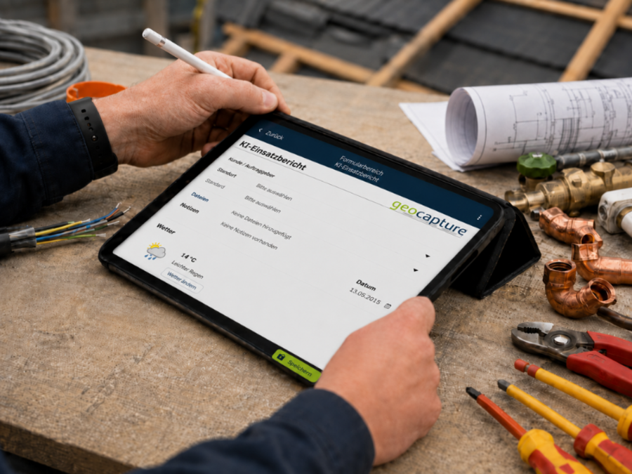Ein Handwerker bedient mit einem Eingabestift ein Tablet, auf dem eine geoCapture-KF-Erfassung geöffnet ist. Auf der Werkbank liegen Baupläne, Werkzeuge, Kabel sowie Kupfer- und Rohrverbindungen.